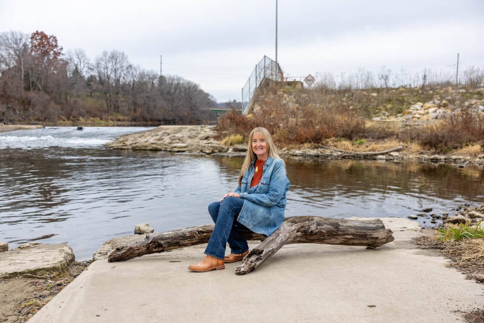 A woman sitting on a log by the river with a bridge in the background.