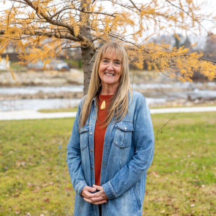 Woman in a denim jacket smiles outdoors with autumn leaves behind her.